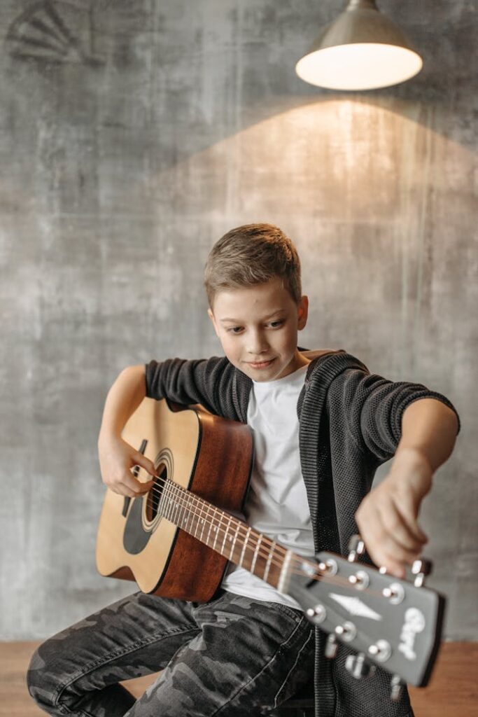 A young boy adjusts his acoustic guitar indoors under warm lighting.